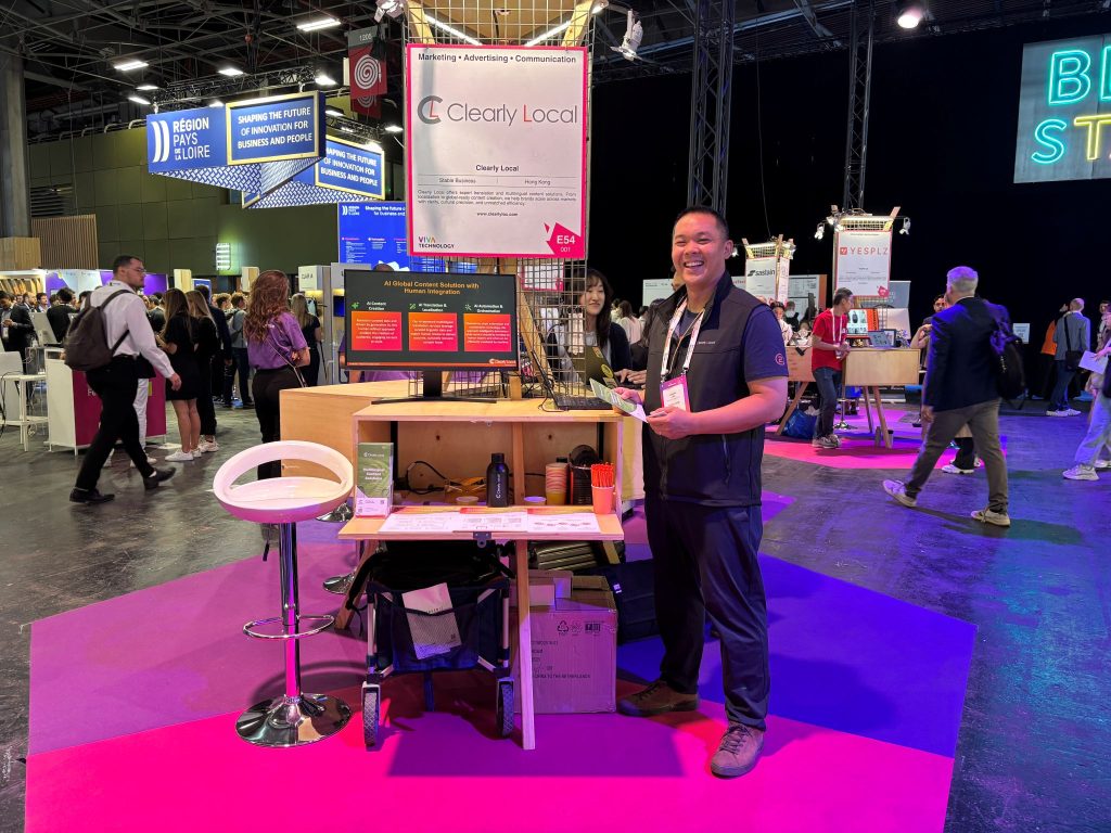 IMG_3119 A smiling man stands in front of a Clearly Local exhibition booth at a tech event, surrounded by people and startup displays. The booth features a screen, promotional materials, and a bright pink and purple floor, with signs and lights from other exhibitors in the background.