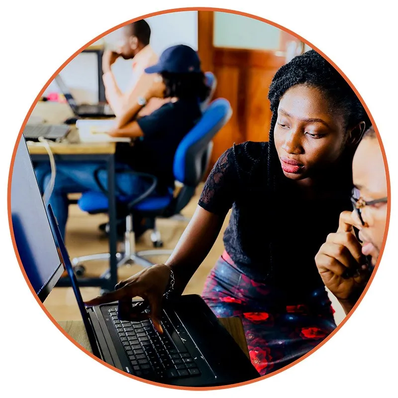 640 A woman is focused, pointing at a laptop screen, guiding a colleague in a vibrant office. Two others work in the background, creating a collaborative atmosphere.