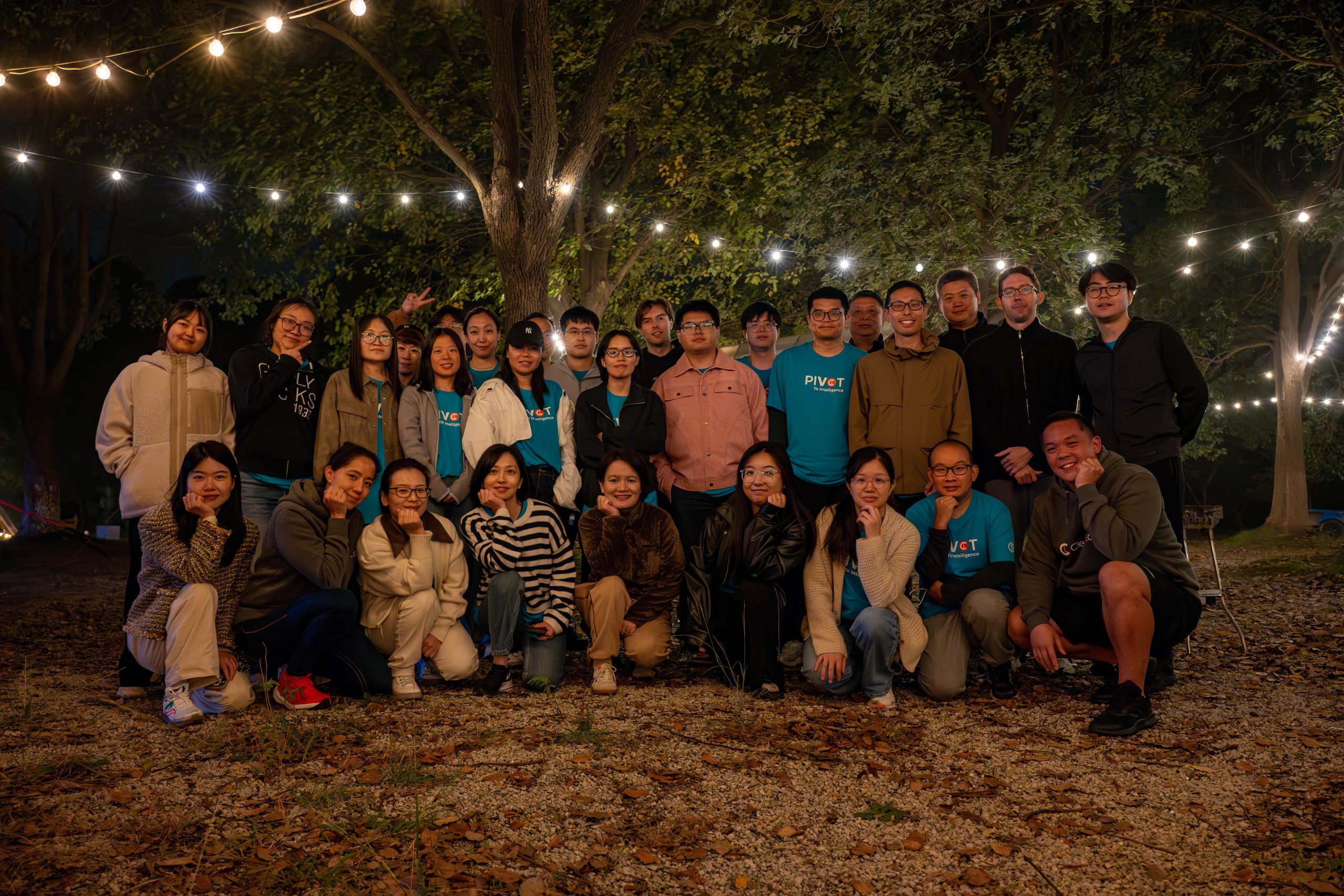 A group of people pose outdoors at night beneath string lights among trees. Most wear casual attire with some in matching blue shirts, conveying a cheerful mood.