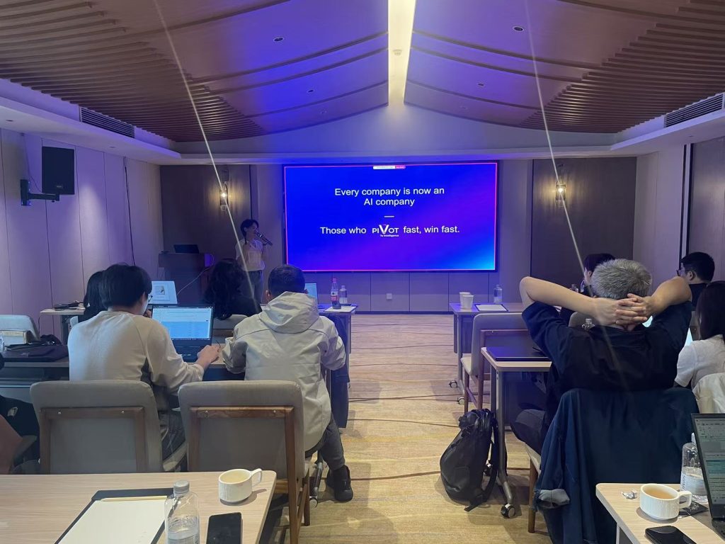 A conference room with attendees seated at tables, facing a presenter. A screen displays: "Every company is now an AI company. Those who pivot fast, win fast." The atmosphere is focused.