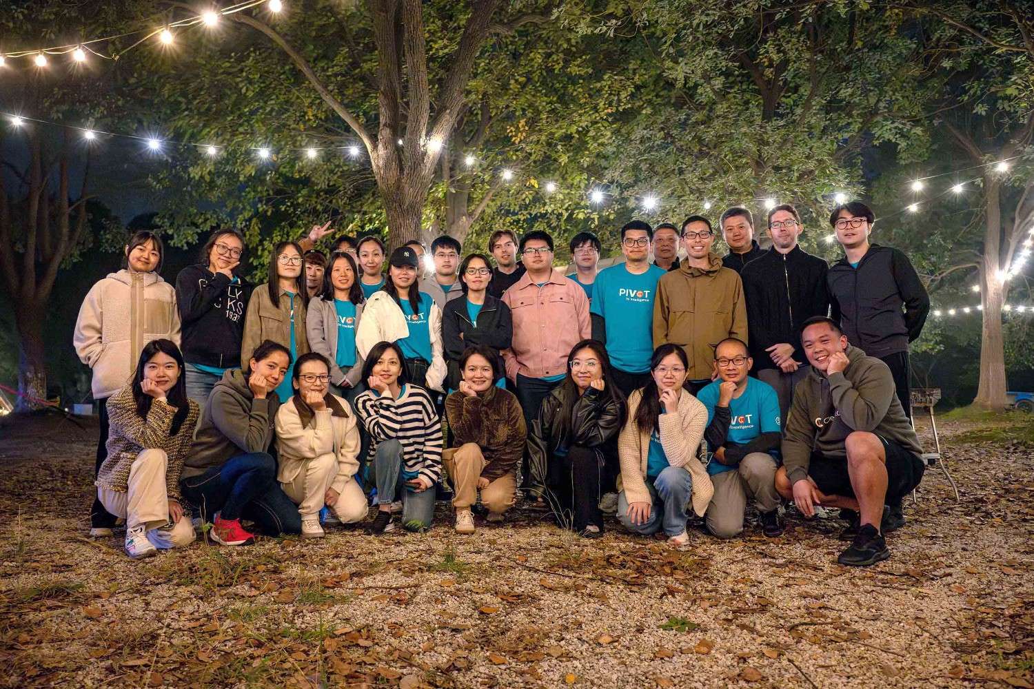 A group of 24 people pose outdoors at night under string lights and trees, smiling warmly, conveying a sense of community and joy.