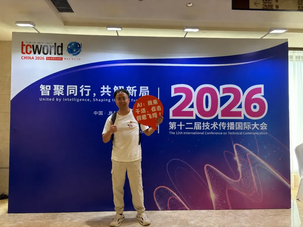 A man stands in front of a large blue-and-white conference backdrop for the 2026 tcworld China event in Shanghai. He is wearing a white T-shirt, light-colored pants, and sneakers, giving a thumbs-up while holding a red speech-bubble sign with Chinese text. The backdrop features prominent “2026” branding, Chinese and English conference text, and abstract wave-like graphics in pink and purple. The photo is taken indoors in a conference venue with overhead lighting.
