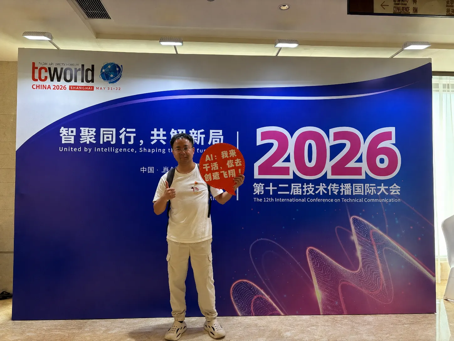 A man stands in front of a large blue-and-white conference backdrop for the 2026 tcworld China event in Shanghai. He is wearing a white T-shirt, light-colored pants, and sneakers, giving a thumbs-up while holding a red speech-bubble sign with Chinese text. The backdrop features prominent “2026” branding, Chinese and English conference text, and abstract wave-like graphics in pink and purple. The photo is taken indoors in a conference venue with overhead lighting.