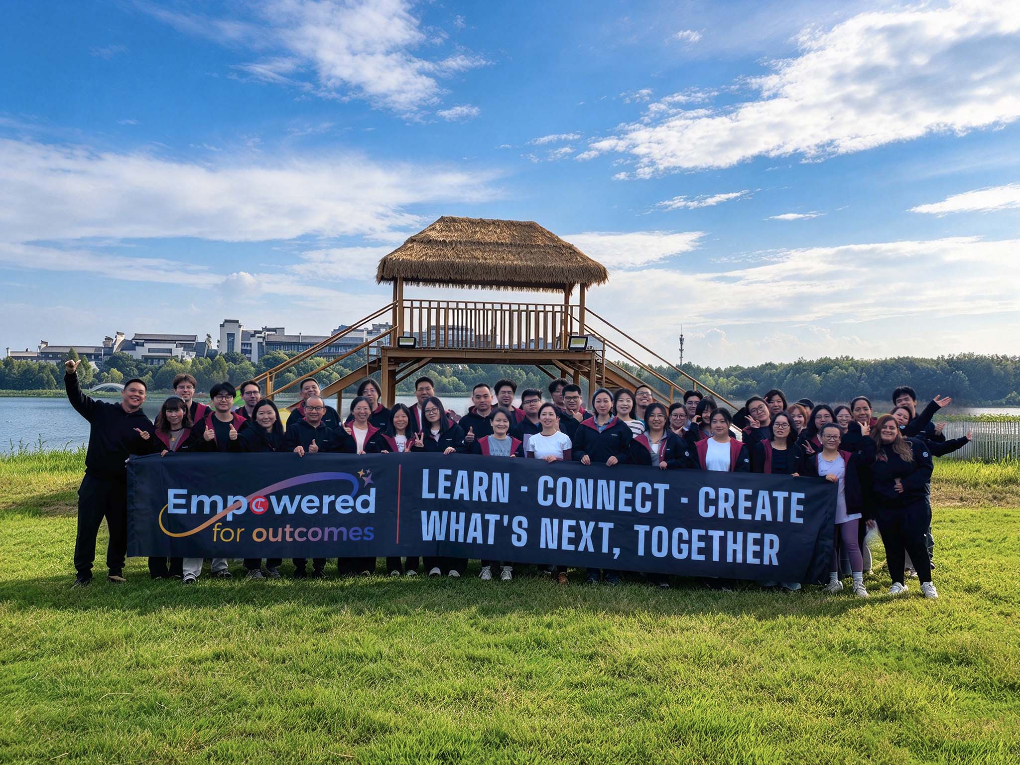 A group of people stands in front of a wooden structure by a lake, holding a banner that reads, "Empowered for outcomes. LEARN * CONNECT * CREATE WHAT'S NEXT, TOGETHER."